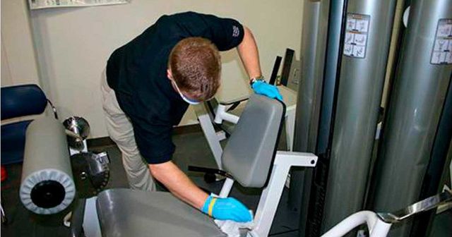 A man wearing a mask and gloves is cleaning a gym machine.