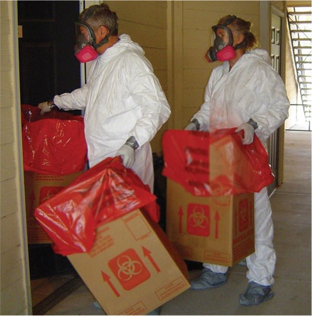 Two people wearing gas masks are carrying boxes with biohazard bags on them