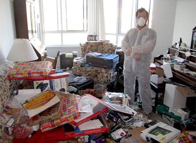 A man wearing a mask is standing in a messy living room.