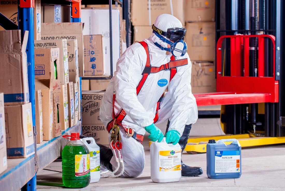 A man in a protective suit is kneeling down in a warehouse with bottles of chemicals.