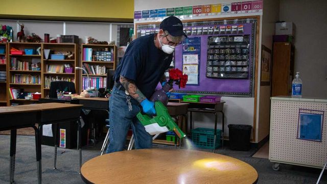 A man wearing a mask is disinfecting a table in a classroom.