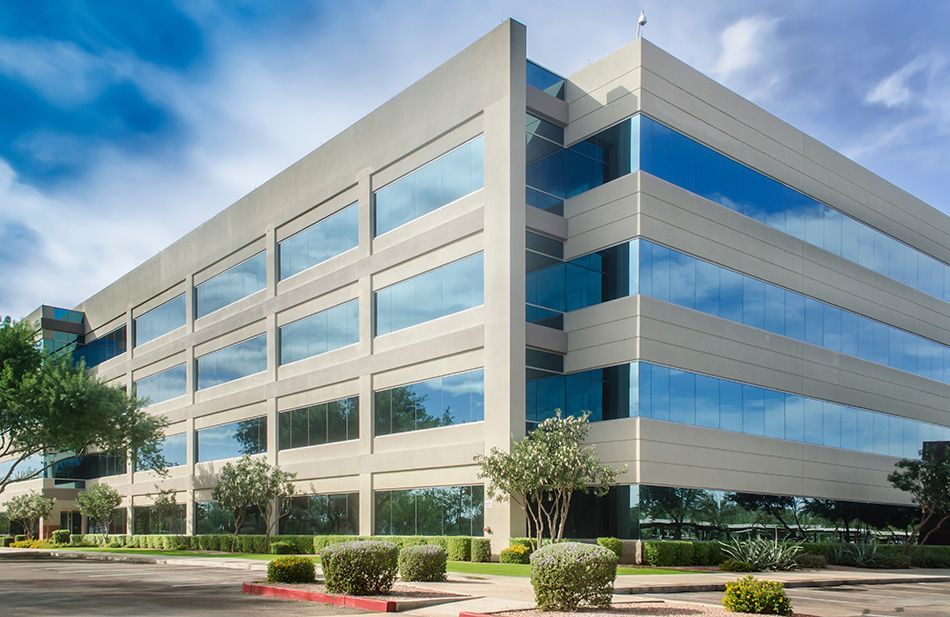 Modern beige office building with large blue-tinted windows reflecting the sky, small bushes, and green grass.