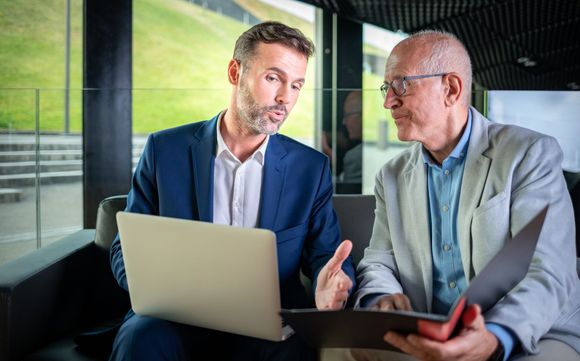 Two men in suits reviewing laptop and documents, gesturing with serious expressions in modern office.