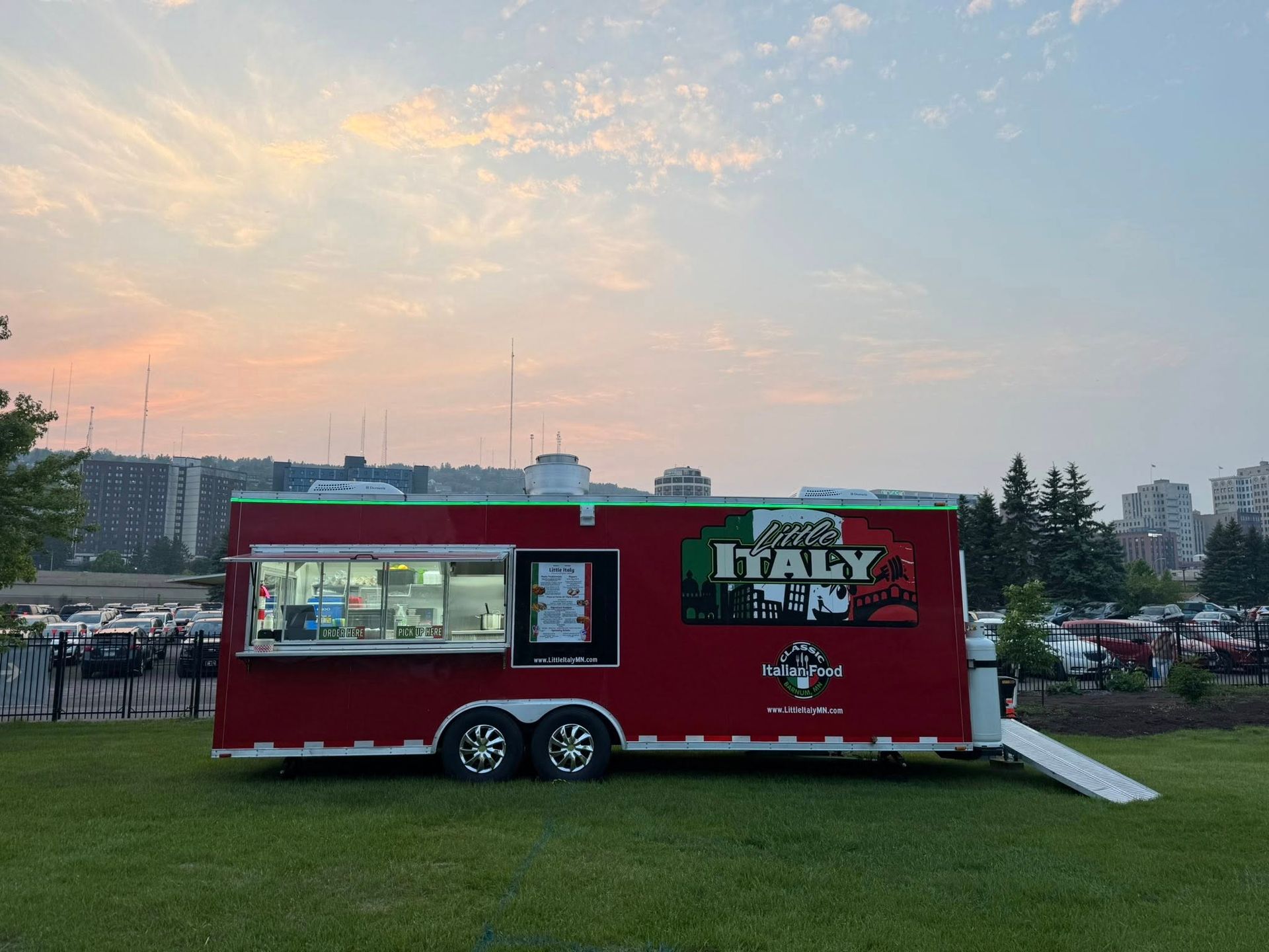 Red Italian food truck with open window on a grassy field under a twilight sky.