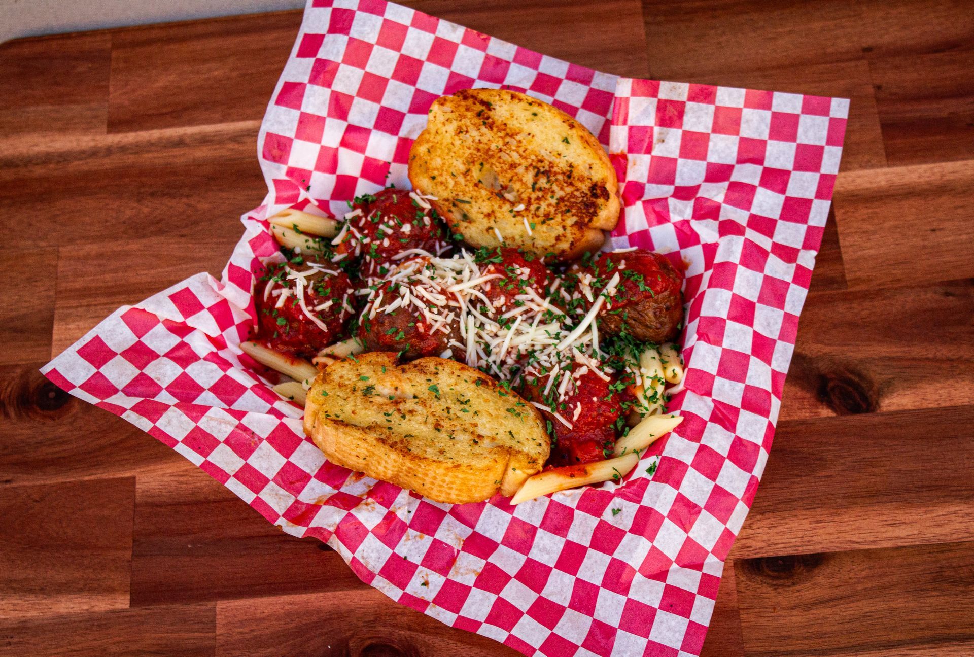Meatballs and pasta with garlic bread, served on a red and white checkered paper, on a wooden table.