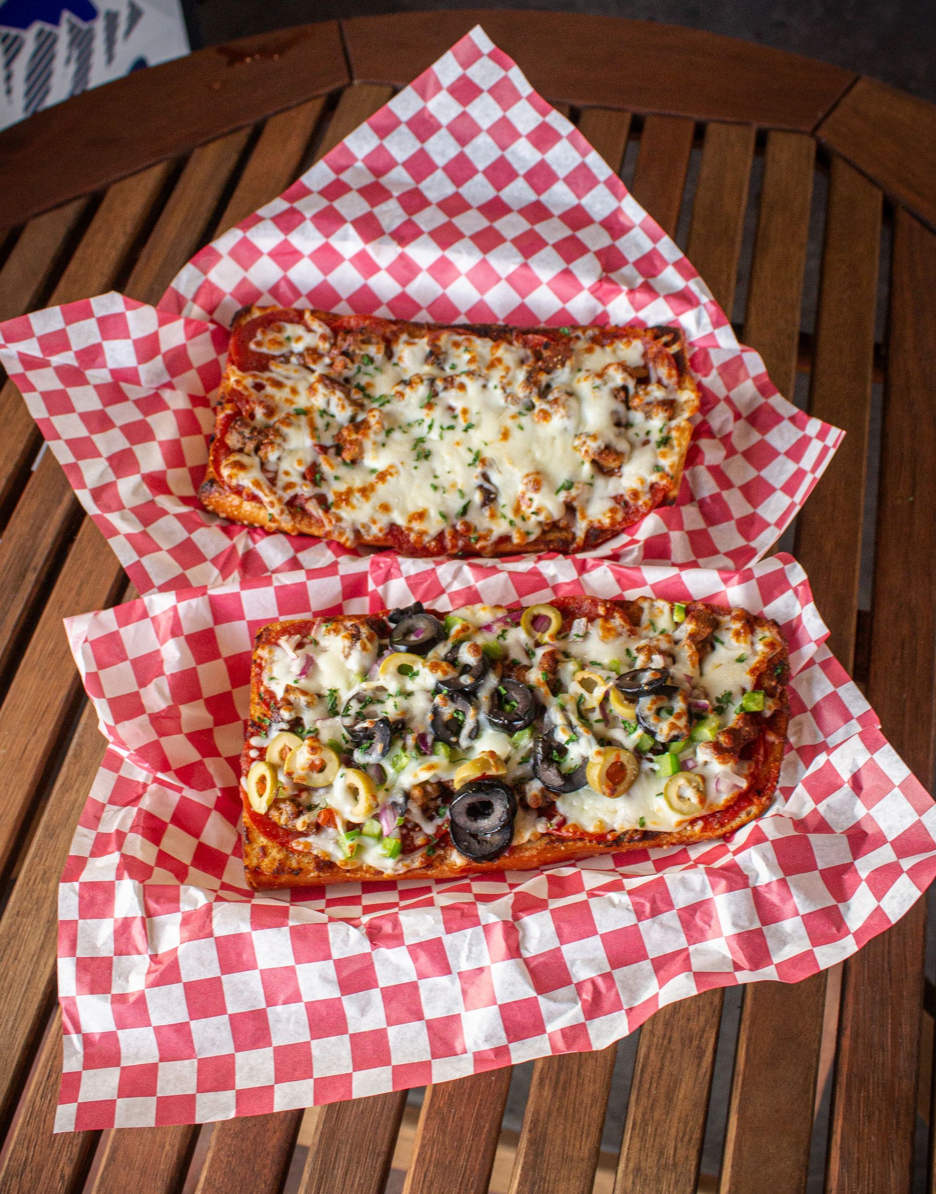 Two pizza bread slices on red checkered paper on a wooden table, one with toppings, the other with cheese.