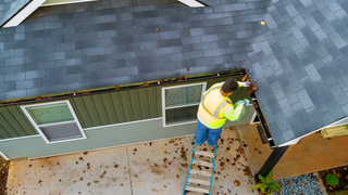 An aerial view of a man on a ladder cleaning a gutter on the side of a house.