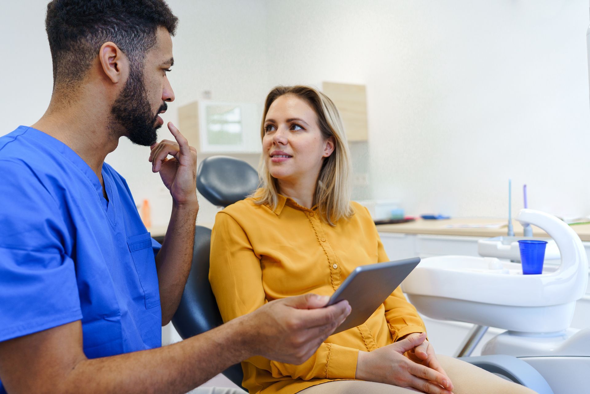 Dental professional consulting with a patient in a clinic, holding a tablet while seated beside dental equipment.