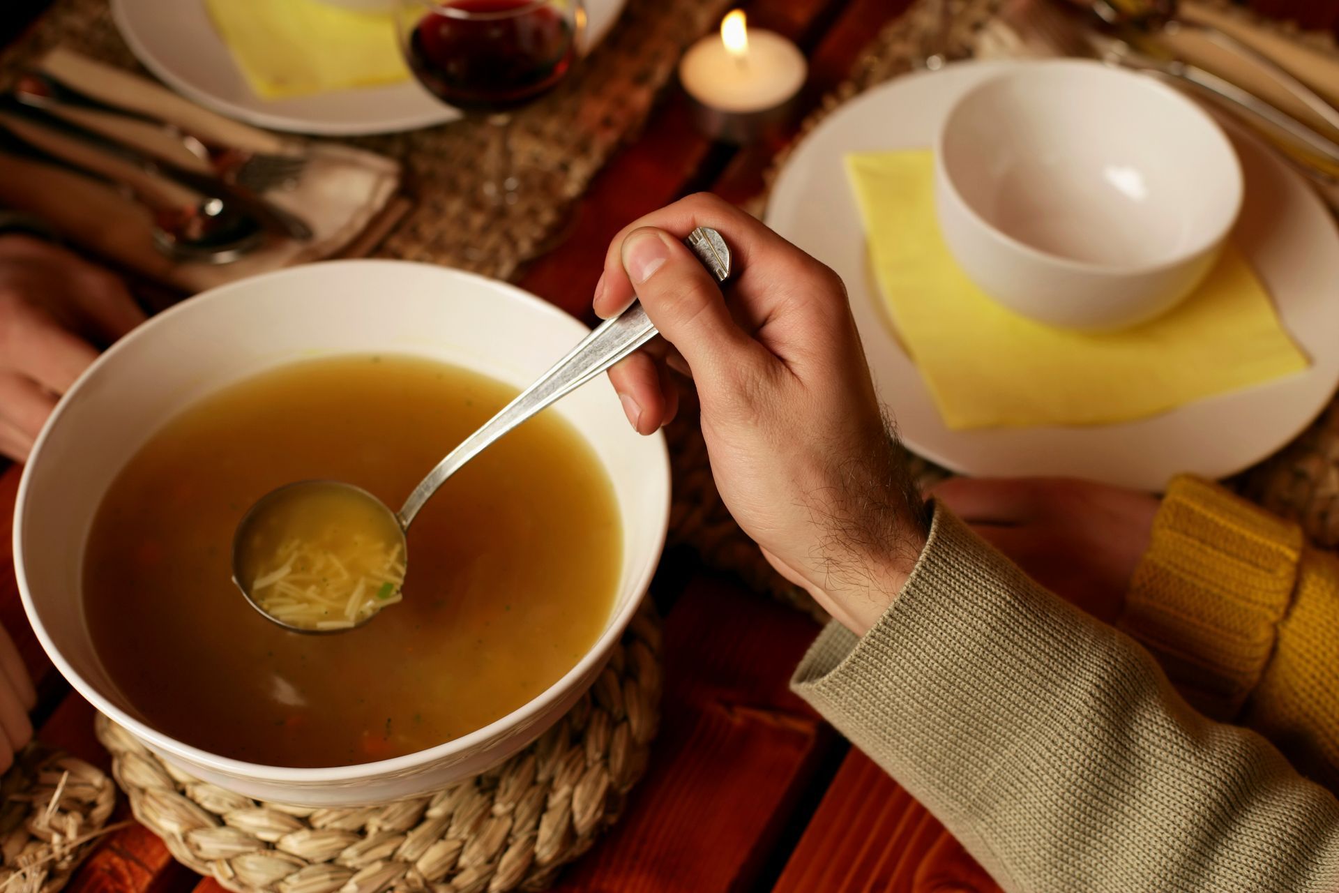 Hand stirring a bowl of warm soup on a table with a cup and candlelight