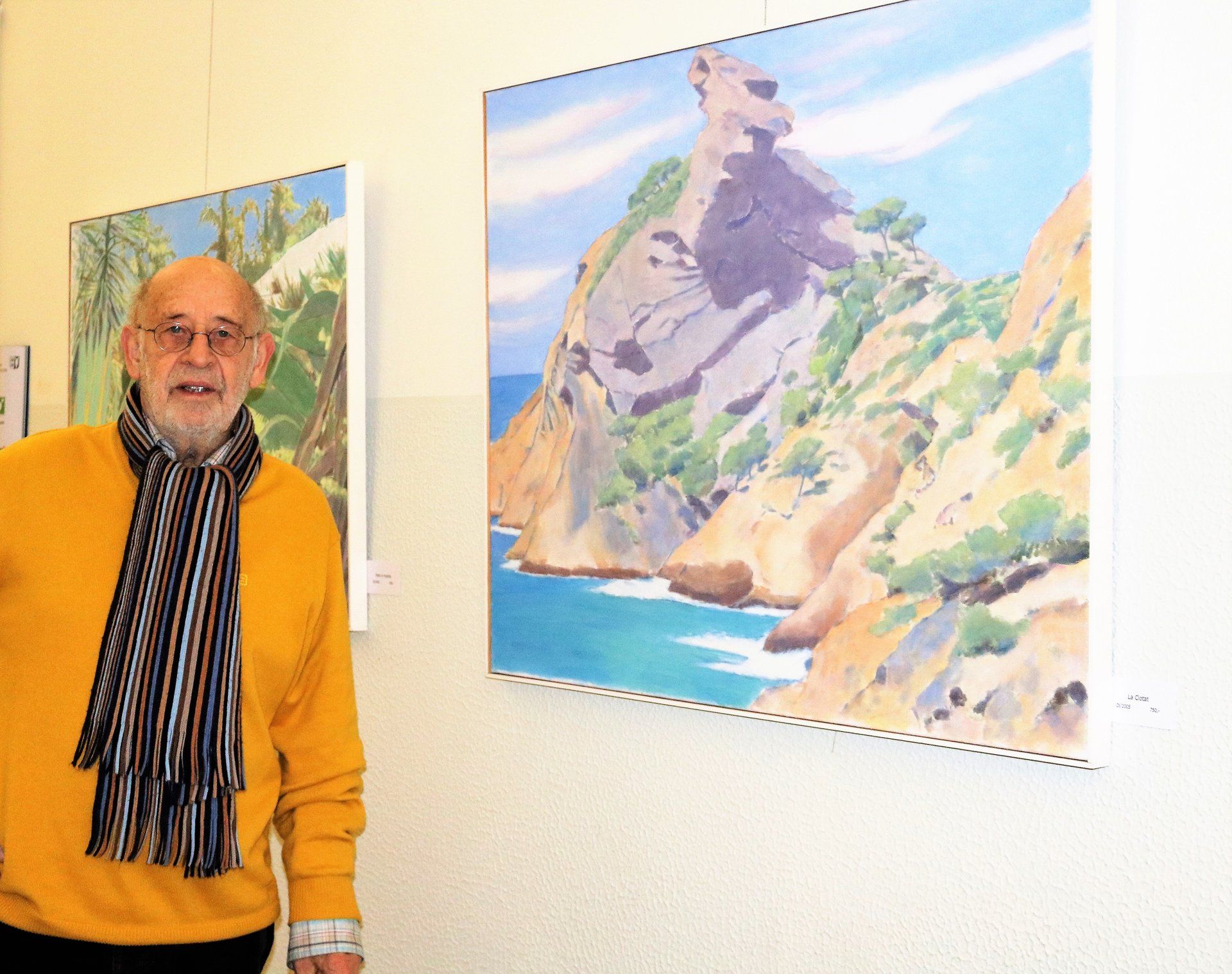 Ein Mann in einem gelben Pullover und Schal steht vor zwei Gemälden an einer Wand.