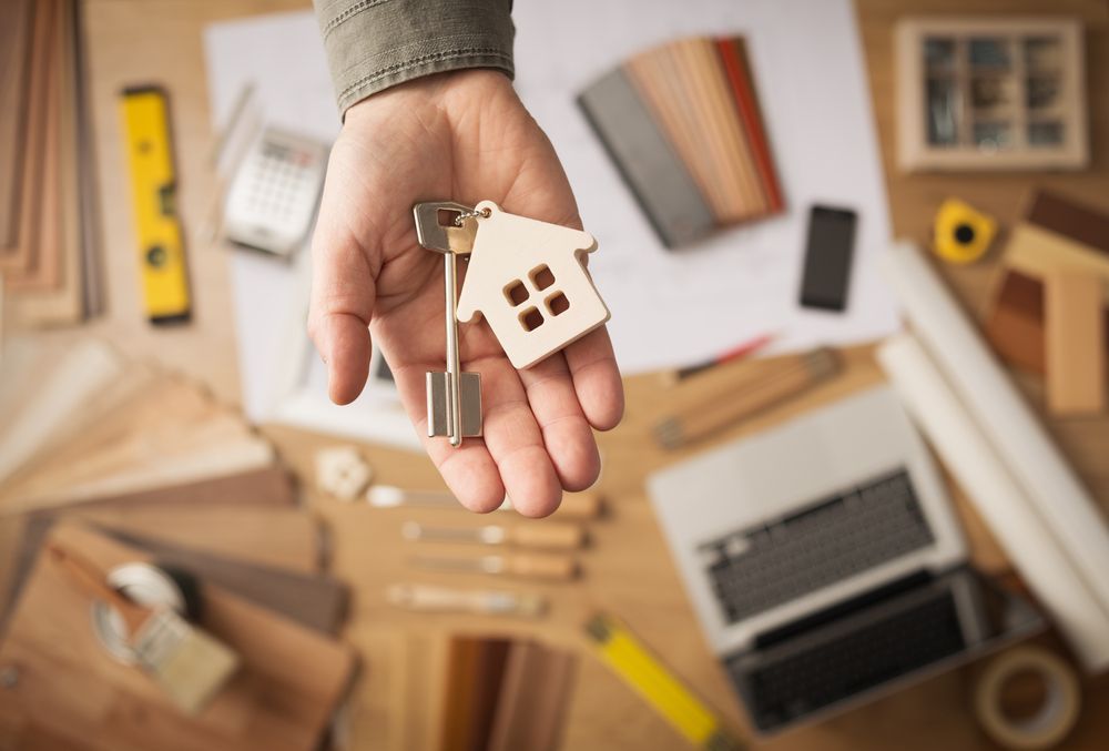 Hand Holding Keys With House-shaped Keychain Above a Desk — Property Transfer Professionals In Bateau Bay, NSW