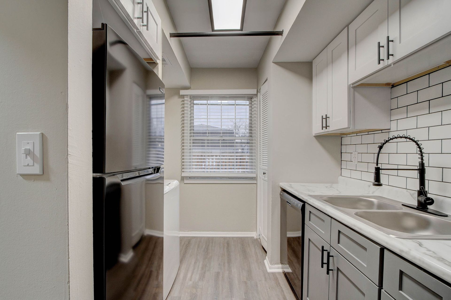 Narrow kitchen with gray and white cabinets, a window, and a black refrigerator.