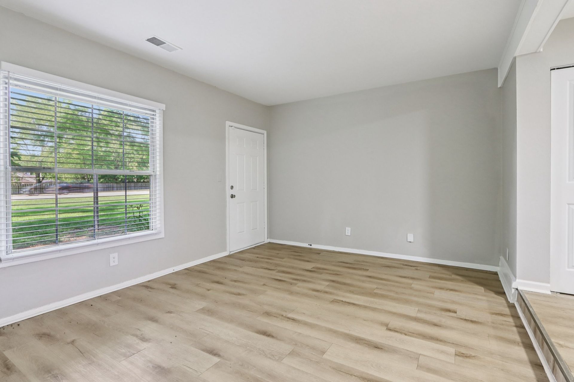 Empty room with gray walls, wood-look floor, and large window with view of trees.
