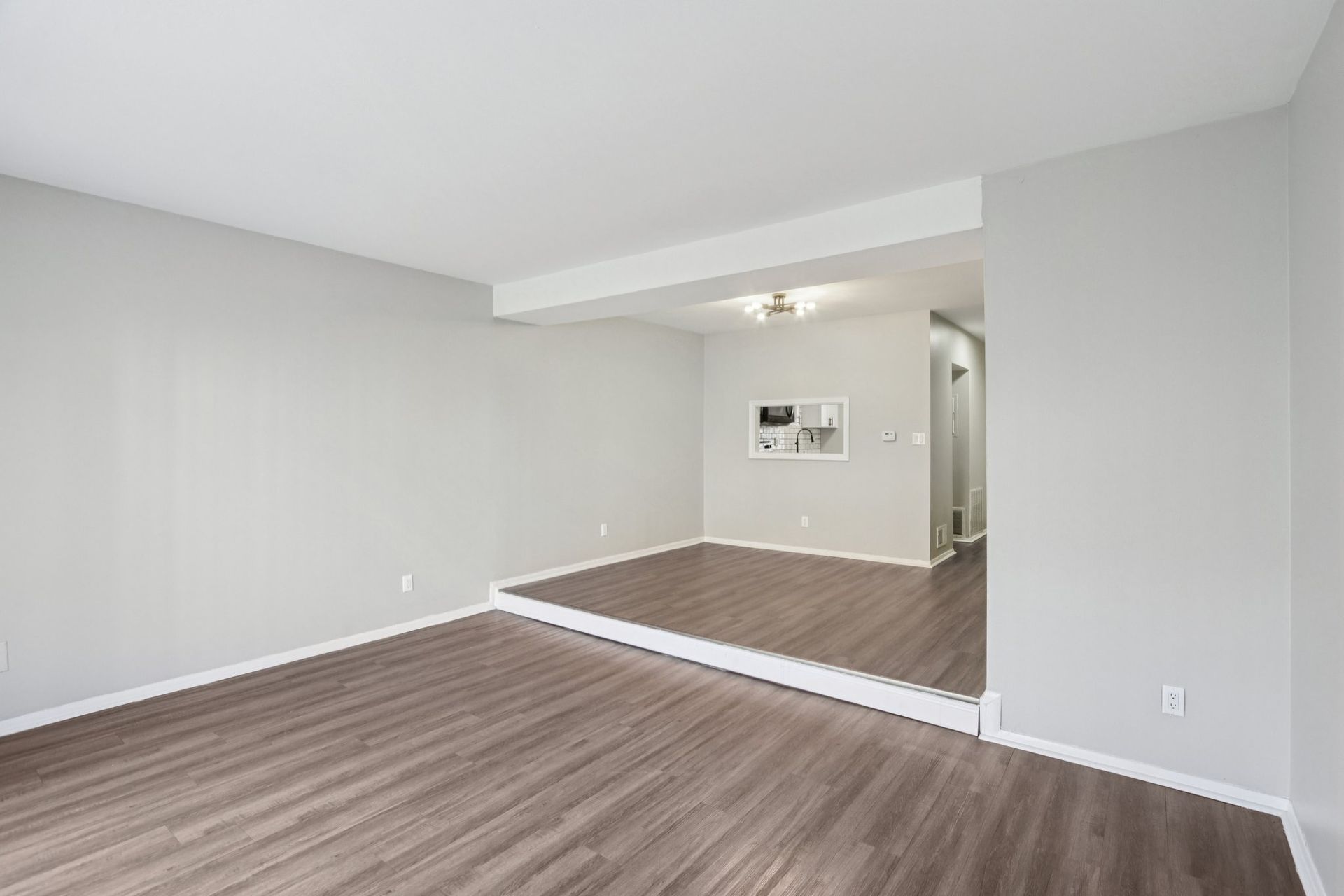Empty living room with gray walls, wood floor, and a raised area with a kitchen.