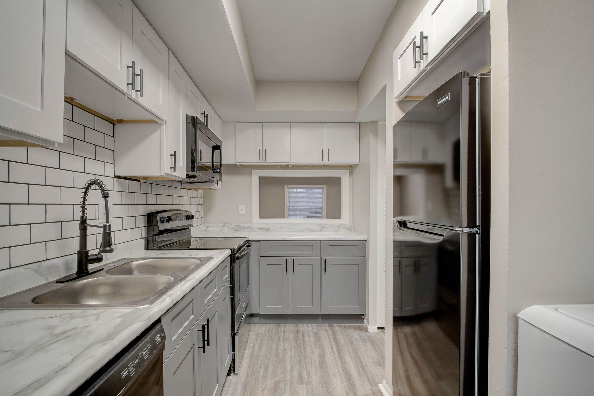 Modern, narrow kitchen with white and gray cabinets, stainless appliances, and a window.