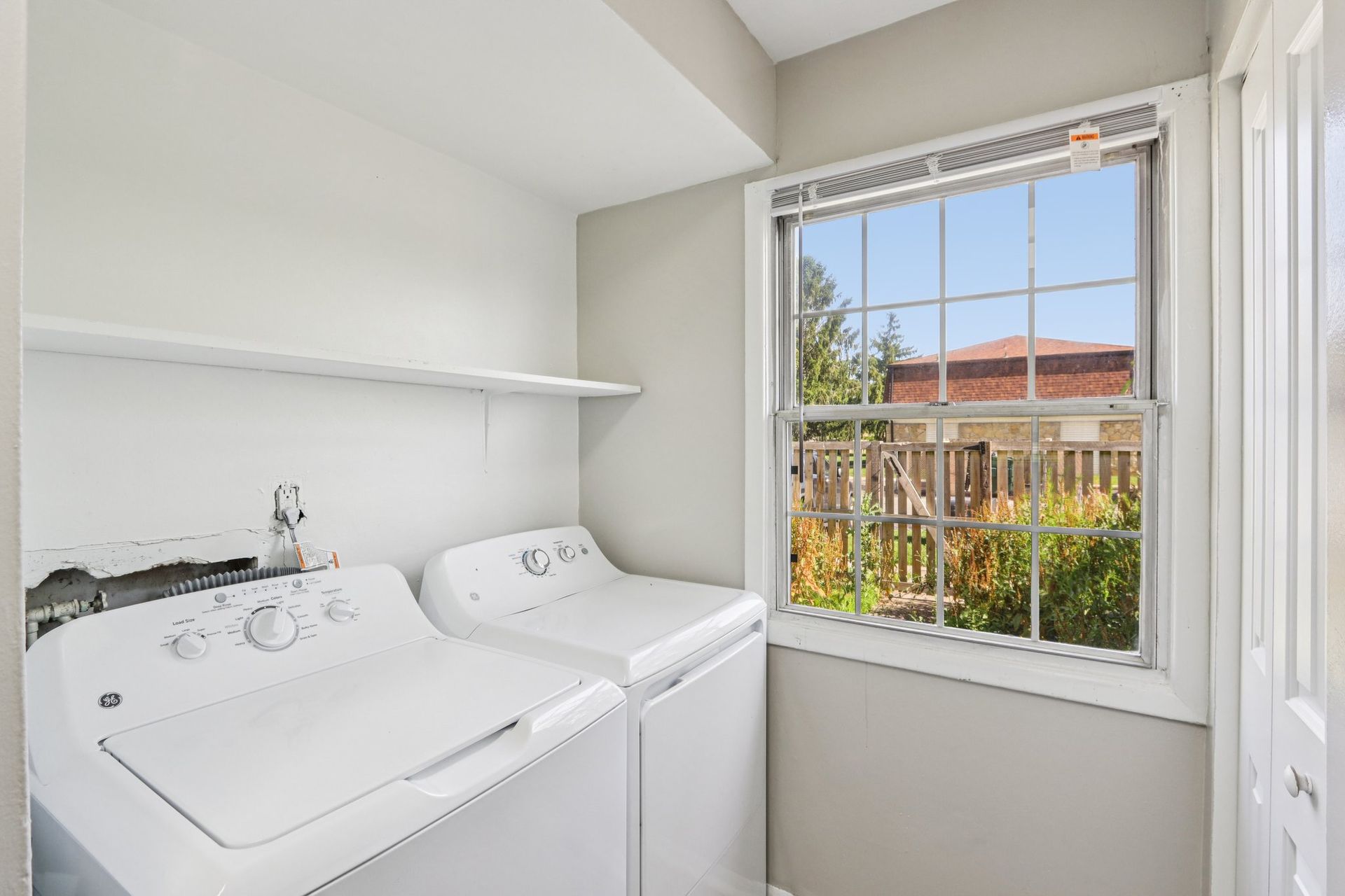 Laundry room with washer, dryer, shelf, and window overlooking a yard.