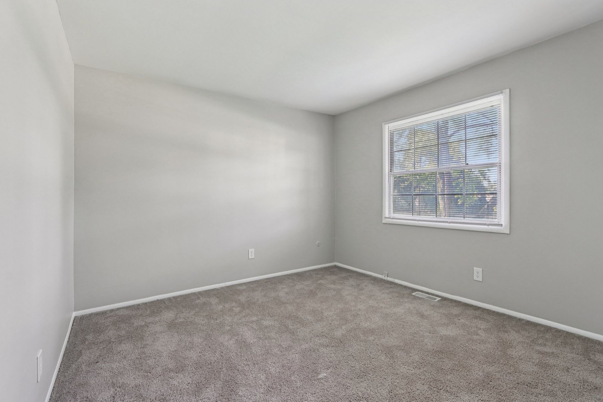 Empty bedroom with gray walls and carpet, a window with blinds, and electrical outlets.