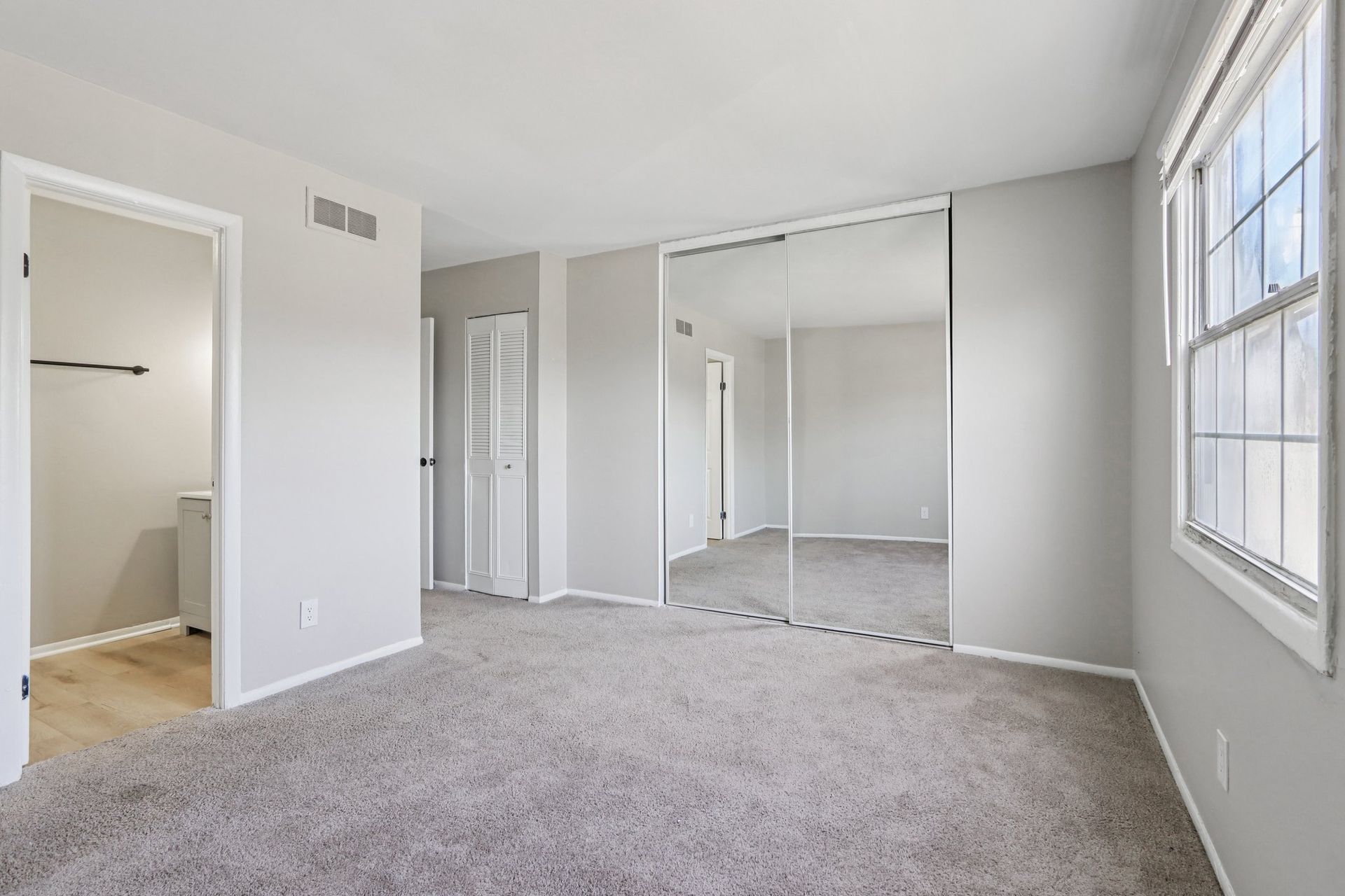 Empty bedroom with gray carpet, mirrored closet doors, and a window.