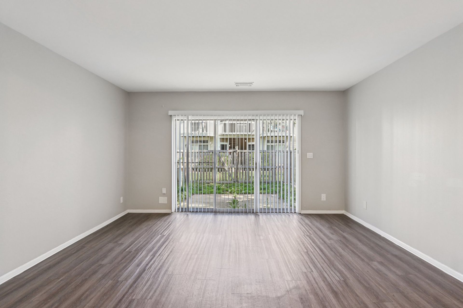 Empty living room with gray walls, wood floors, and a sliding door leading outside.