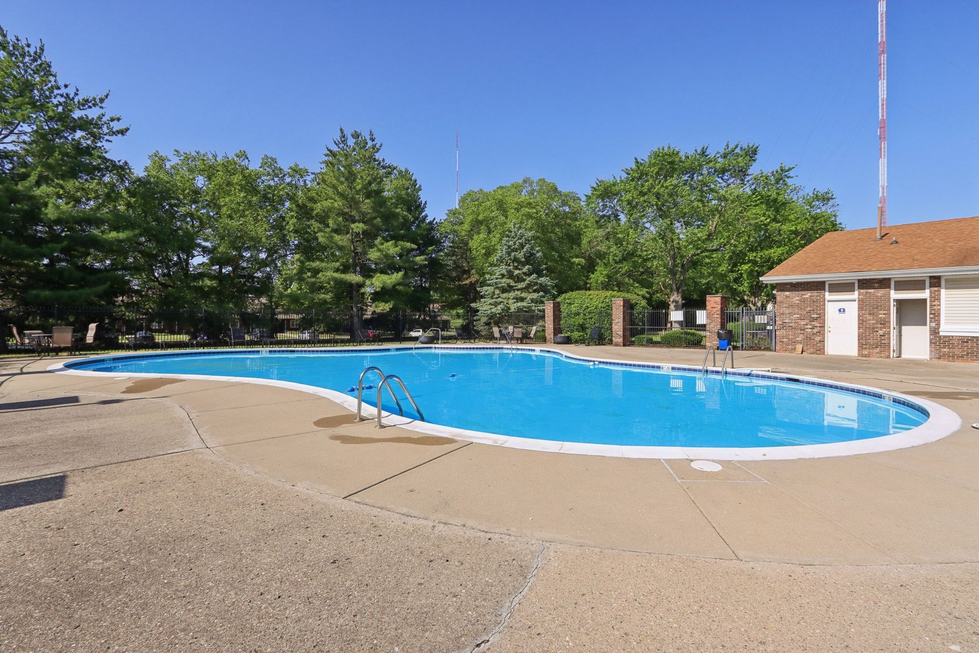 A blue swimming pool, surrounded by concrete and trees, under a clear sky. A small building is to the right.