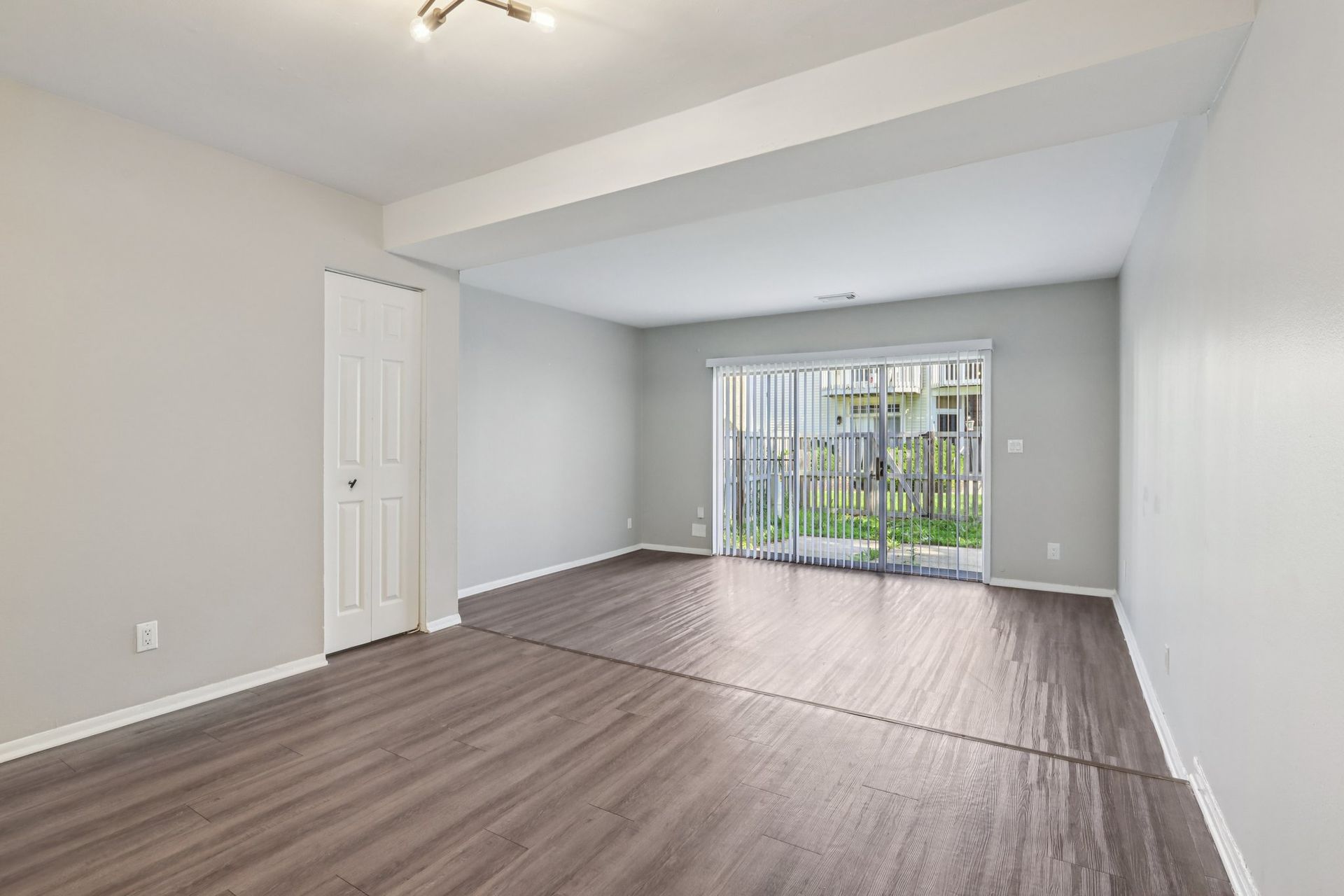 Empty living room with gray walls, dark wood floors, and sliding glass door to backyard.