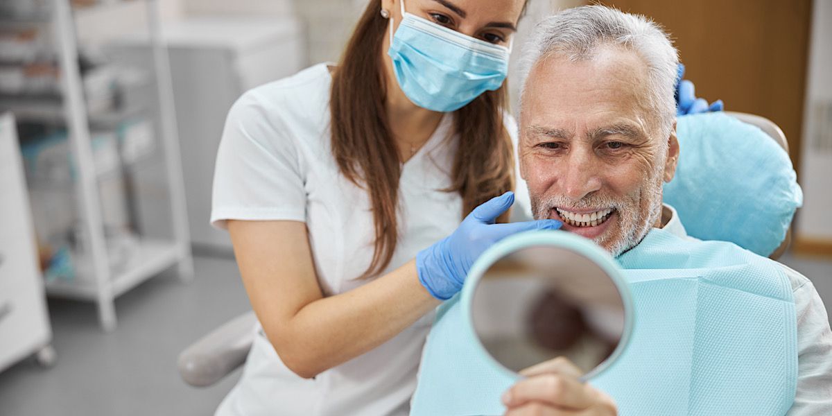 Elderly man sitting in a dental chair next to dental hygienist who is showing him his new smile after an implant placement.