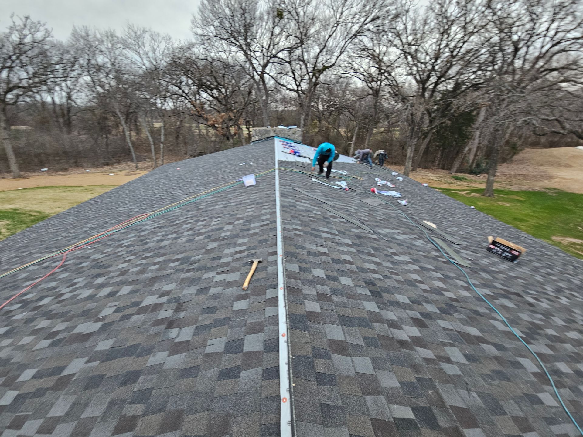 Roofers working on a gray shingle roof on a cloudy day. Trees and grass in the background.