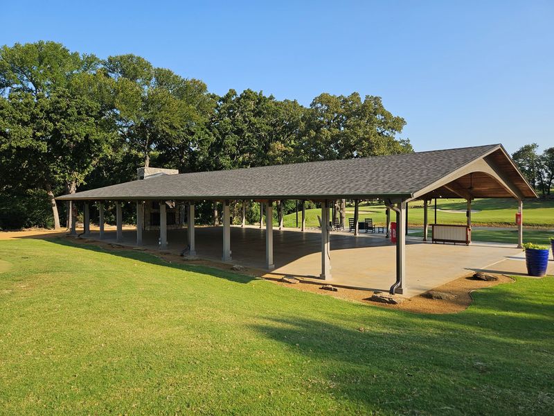 A long, open-air pavilion in a park with a green lawn, a blue trash can, and trees in the background.