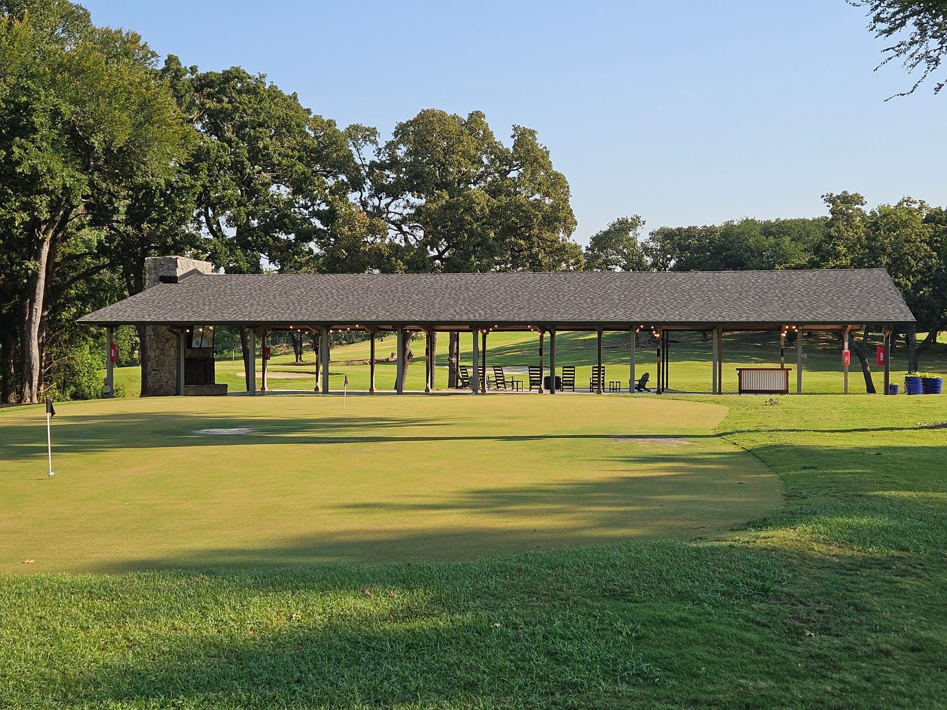 Pavilion with a wood shingle roof over a green grassy area. Trees in the background.