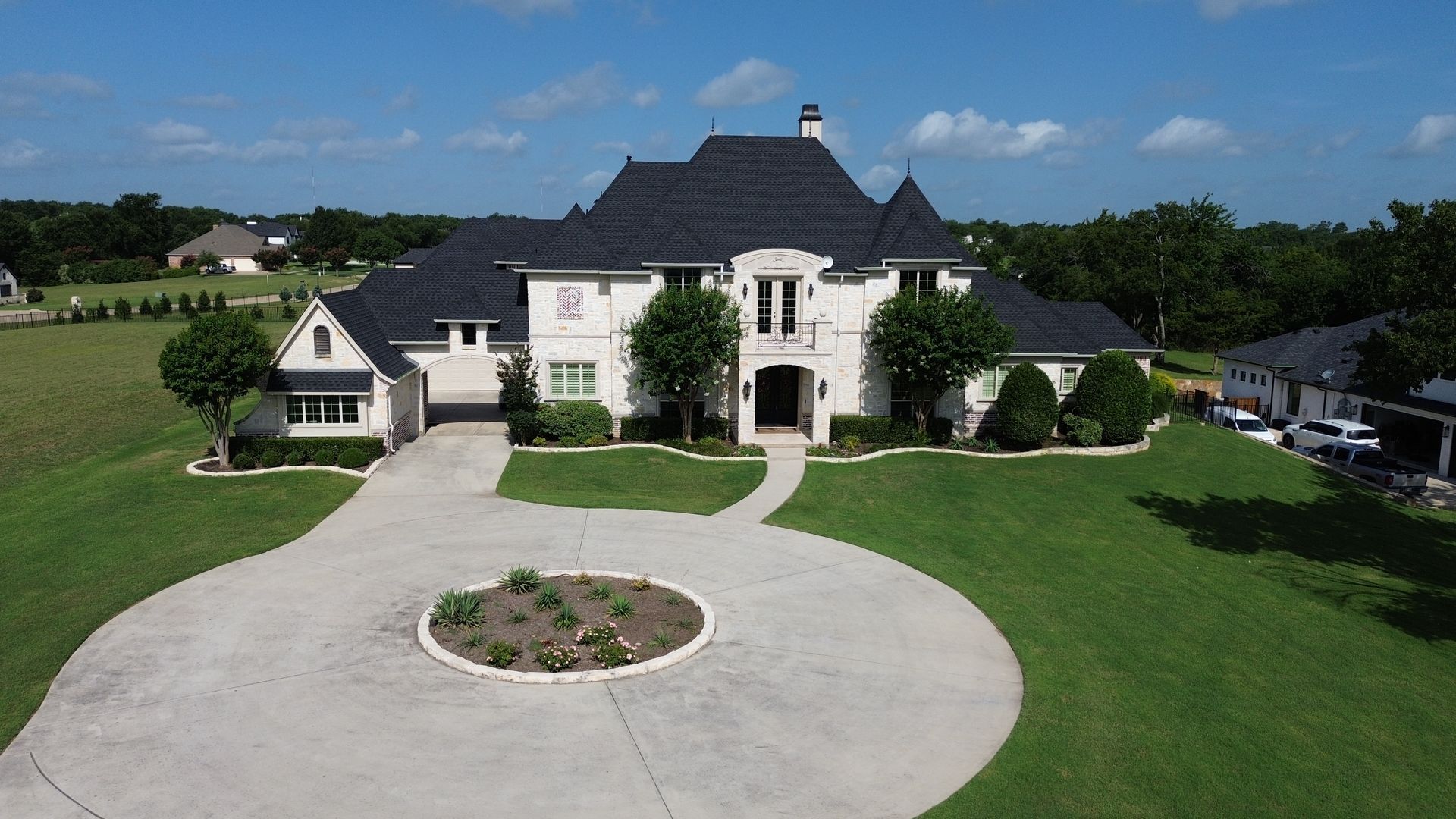Large white house with black roof, circular driveway, and lush green lawn under a blue sky.