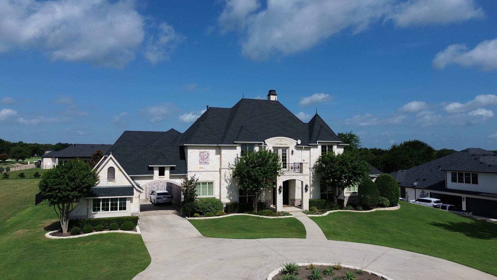 Large white house with dark roof and circular driveway on a grassy hill under blue sky and clouds.