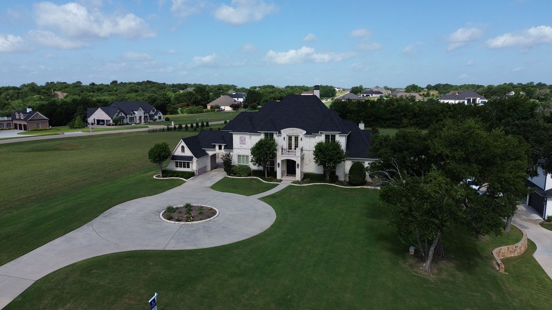 Large, white house with black roof, curved driveway, and green lawn on a sunny day.