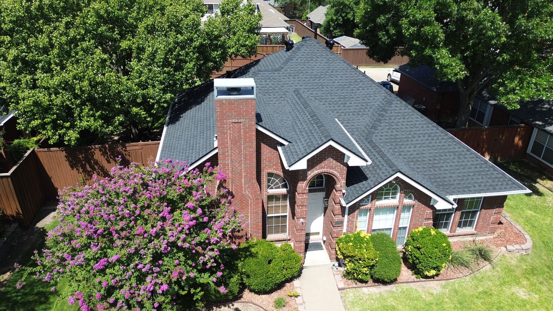Brick house with a dark gray roof, chimney, and front yard with bushes and a tree.