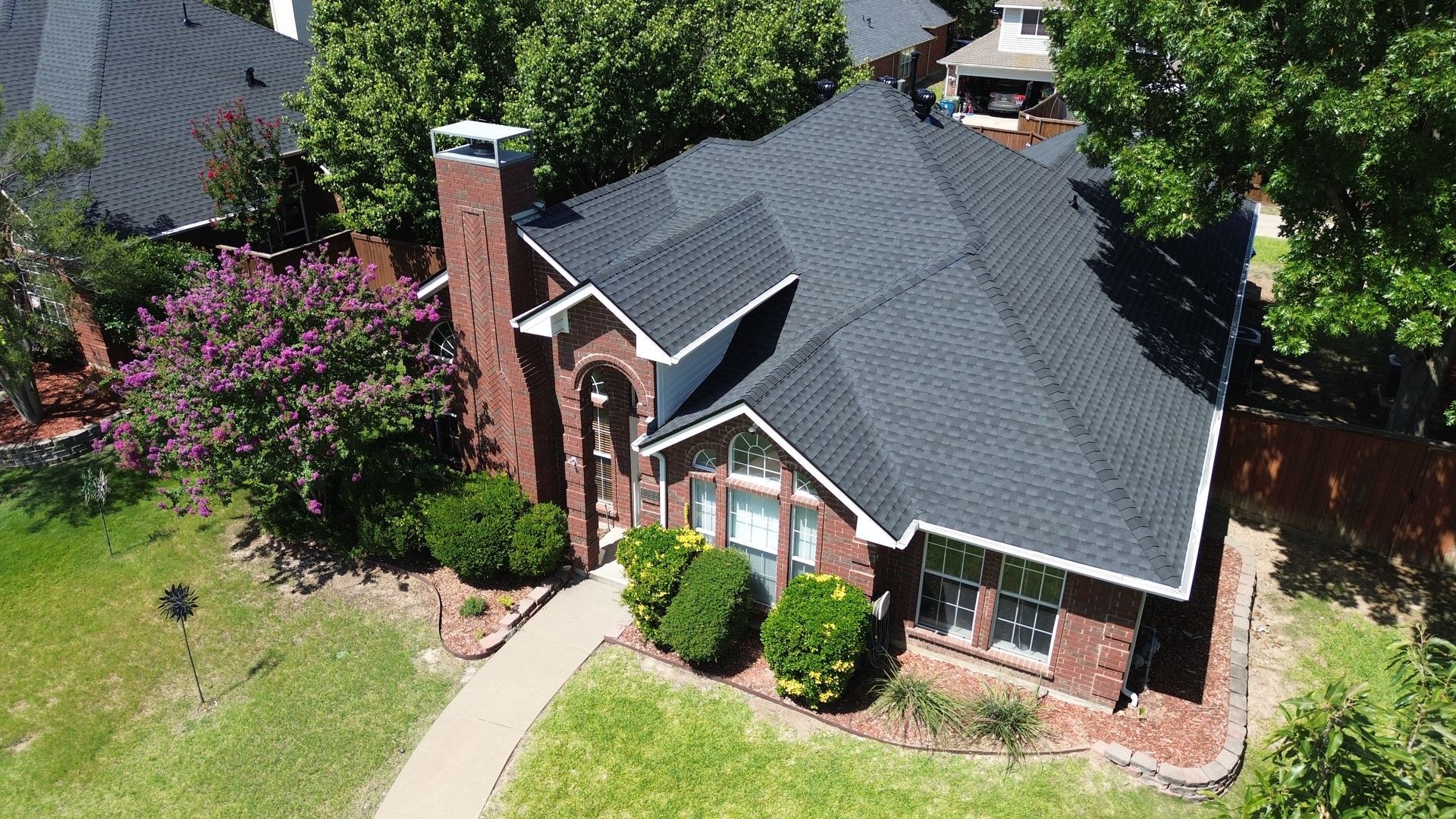 A brick house with a black roof, chimney, and walkway surrounded by green trees and grass.