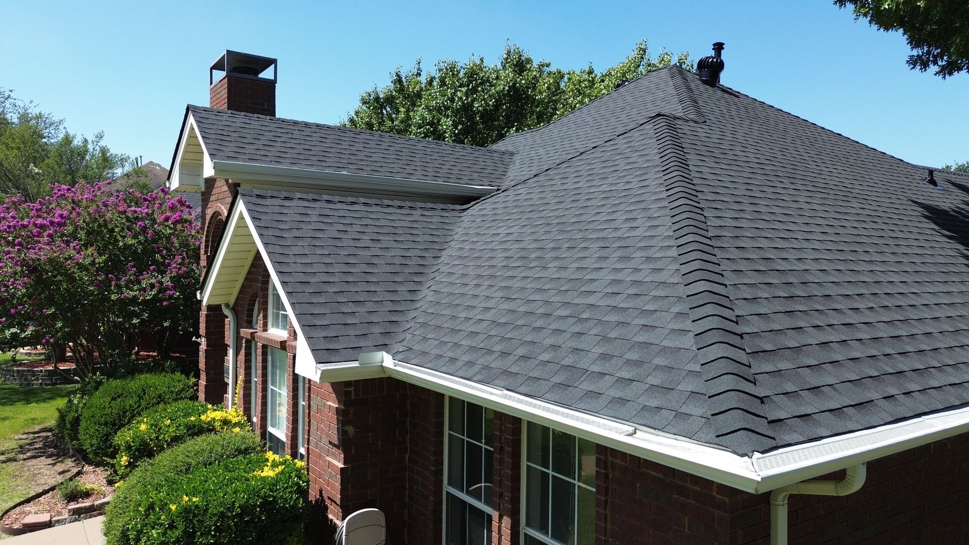 Black shingle roof on a brick house with white trim and a chimney, under a blue sky.