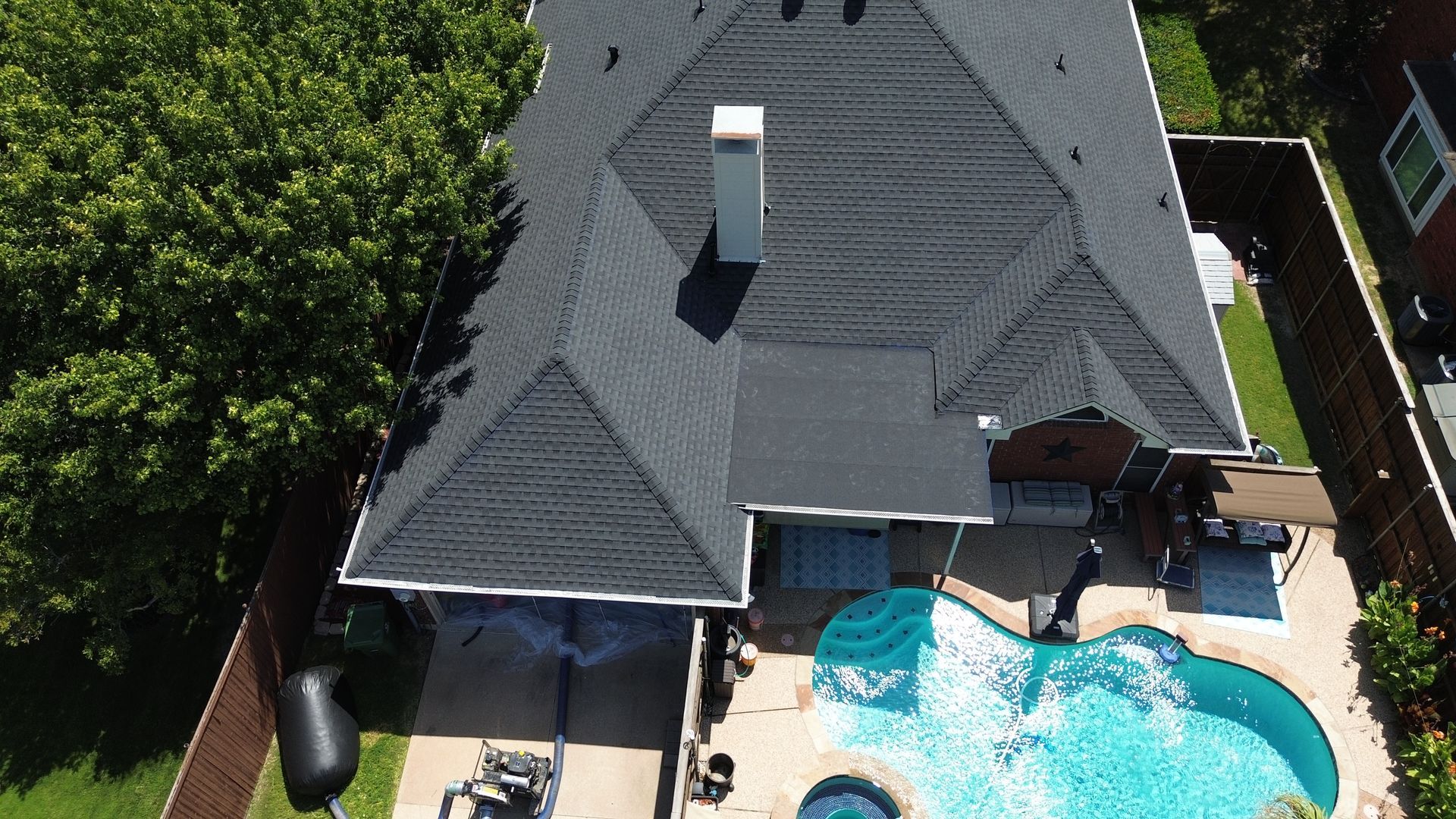 Aerial view of a house with a dark roof, a swimming pool, and a backyard with green trees.