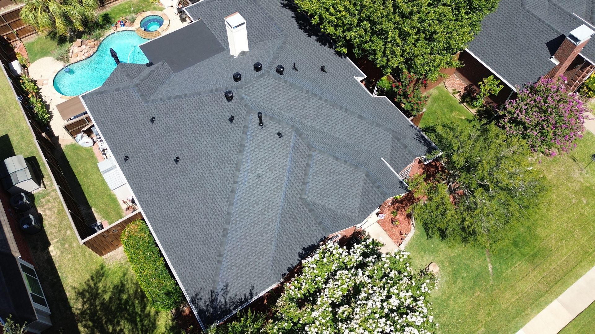 Aerial view of a house with a dark roof, chimney, pool, and surrounding greenery.
