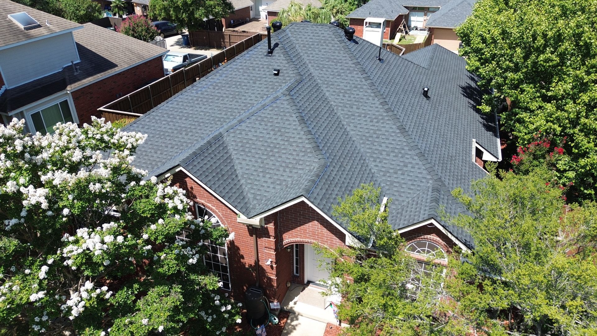 Aerial view of a house with a dark gray shingled roof, red brick, and surrounding green trees.