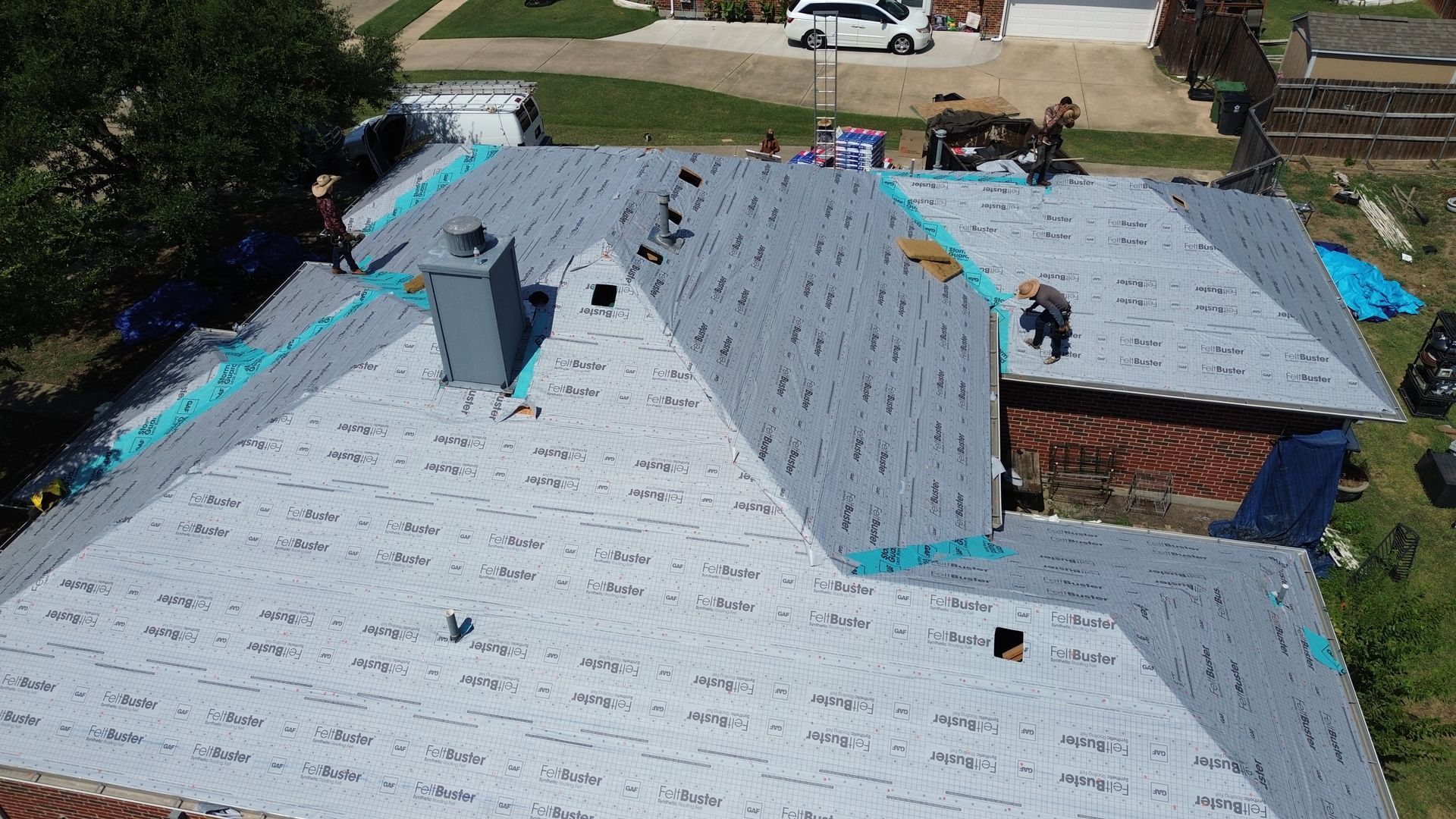 Roofers installing new shingles on a house roof. Blue tarps, gray shingles, sunny day.