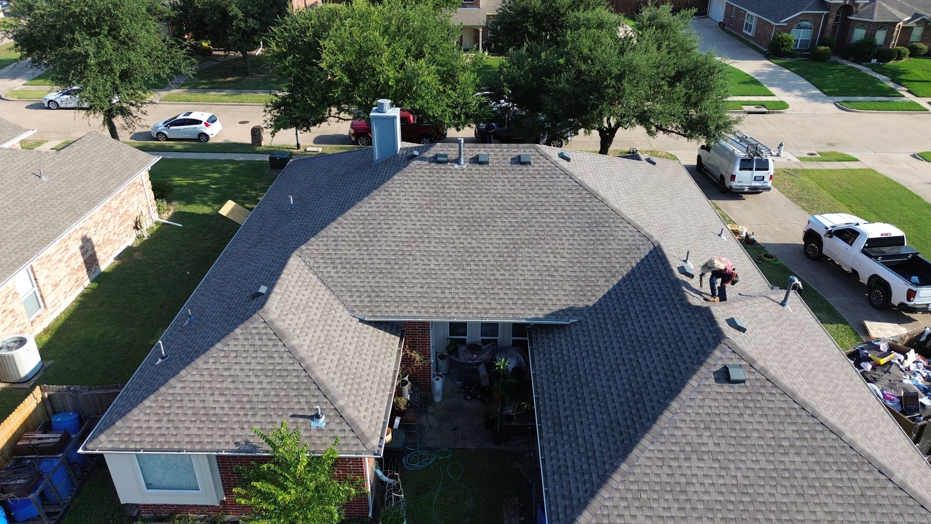 Aerial view of a house with a gray roof. Workers on the roof, vehicles in the driveway. Suburban setting.