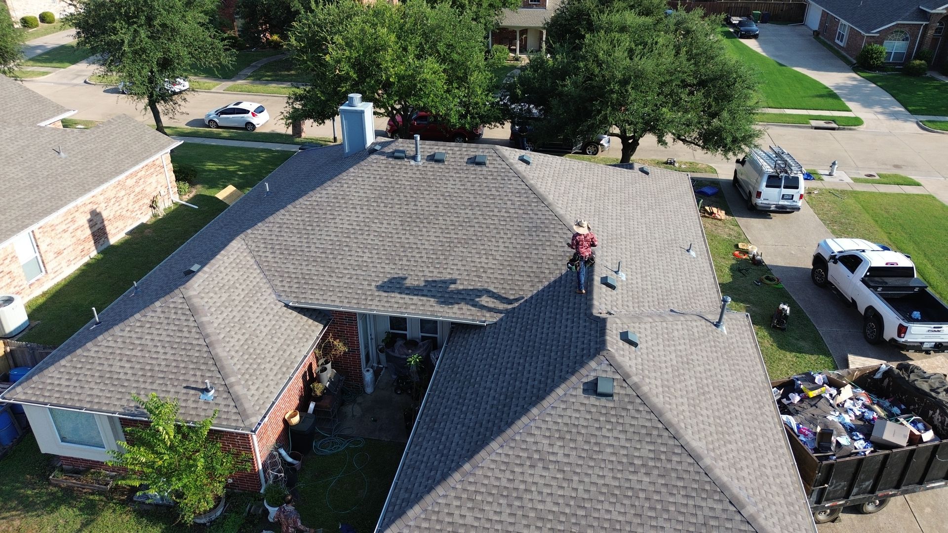 Aerial view of a house roof with a worker, vehicles, and debris bin, likely a roof replacement.
