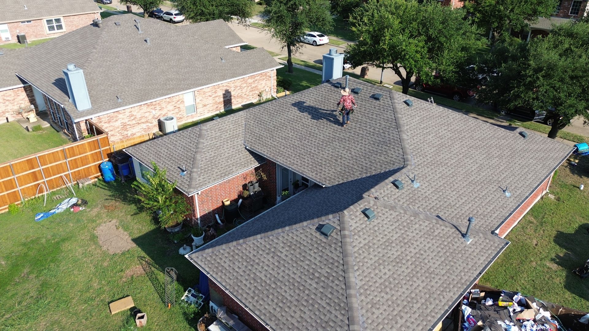 A person working on a shingled roof of a brick house in a residential area, sunny day.