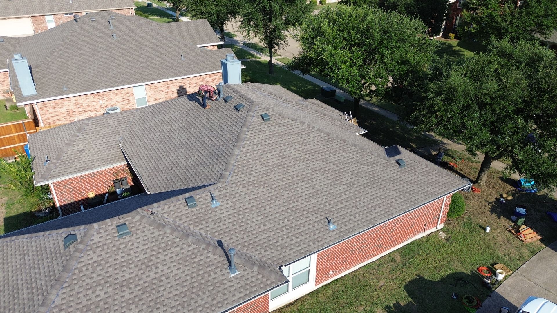 Overhead view of a brown shingle roof with workers, brick house, and green trees.