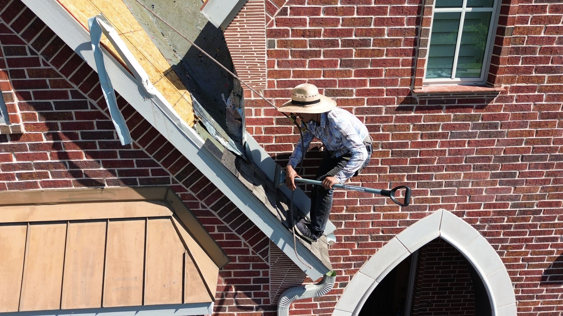 Person on a roof repairs gutter, red brick building in the background.