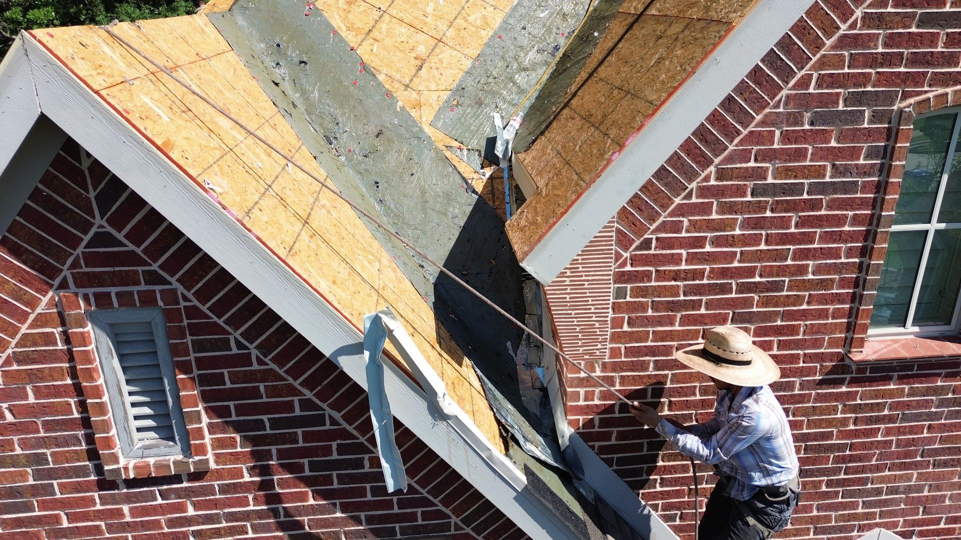 Roofer in a straw hat works on a brick building's roof, partially covered with shingles.