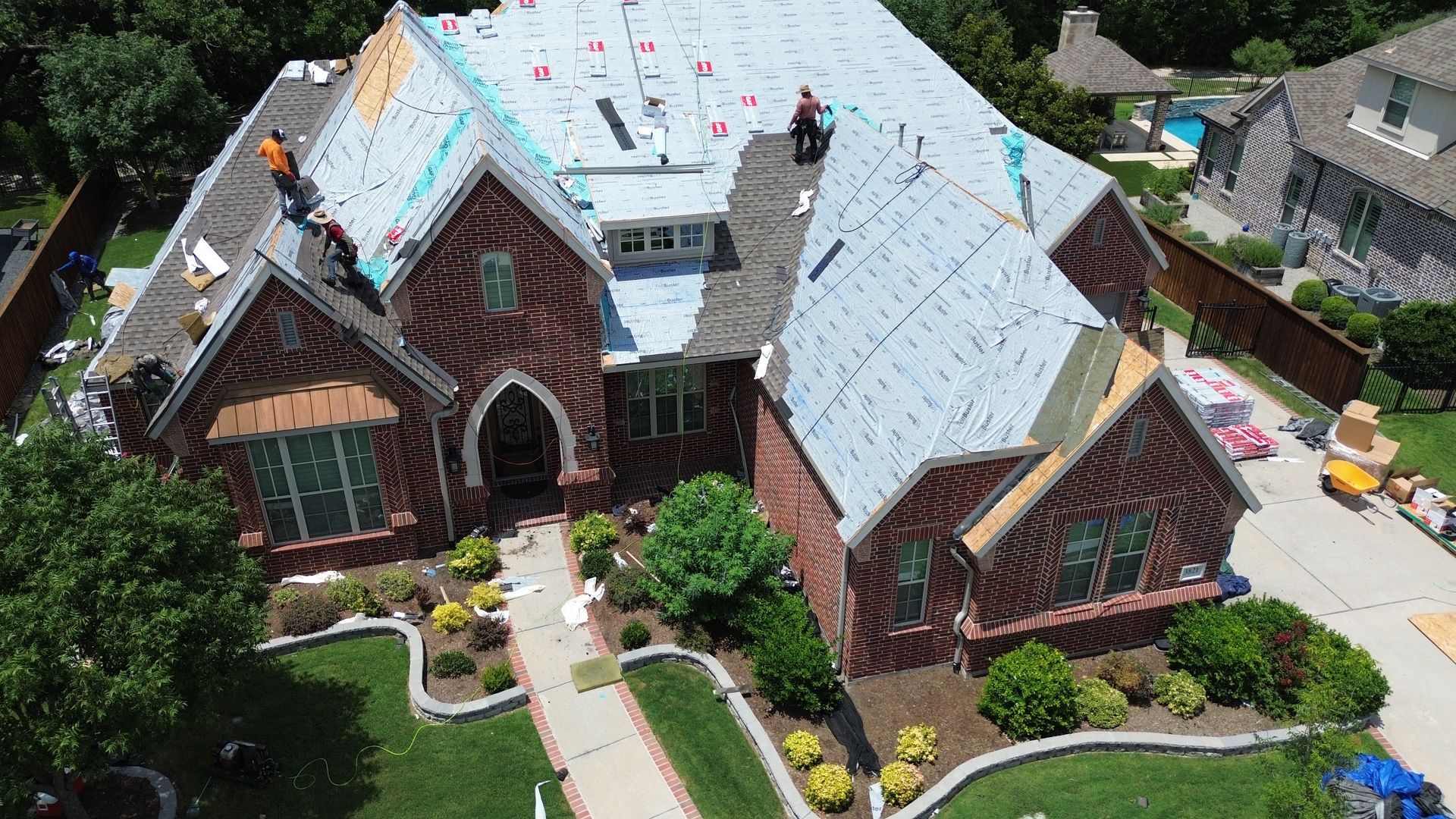 Roofers working on a red brick house with gray roof underlayment. Green lawn, sunny day.