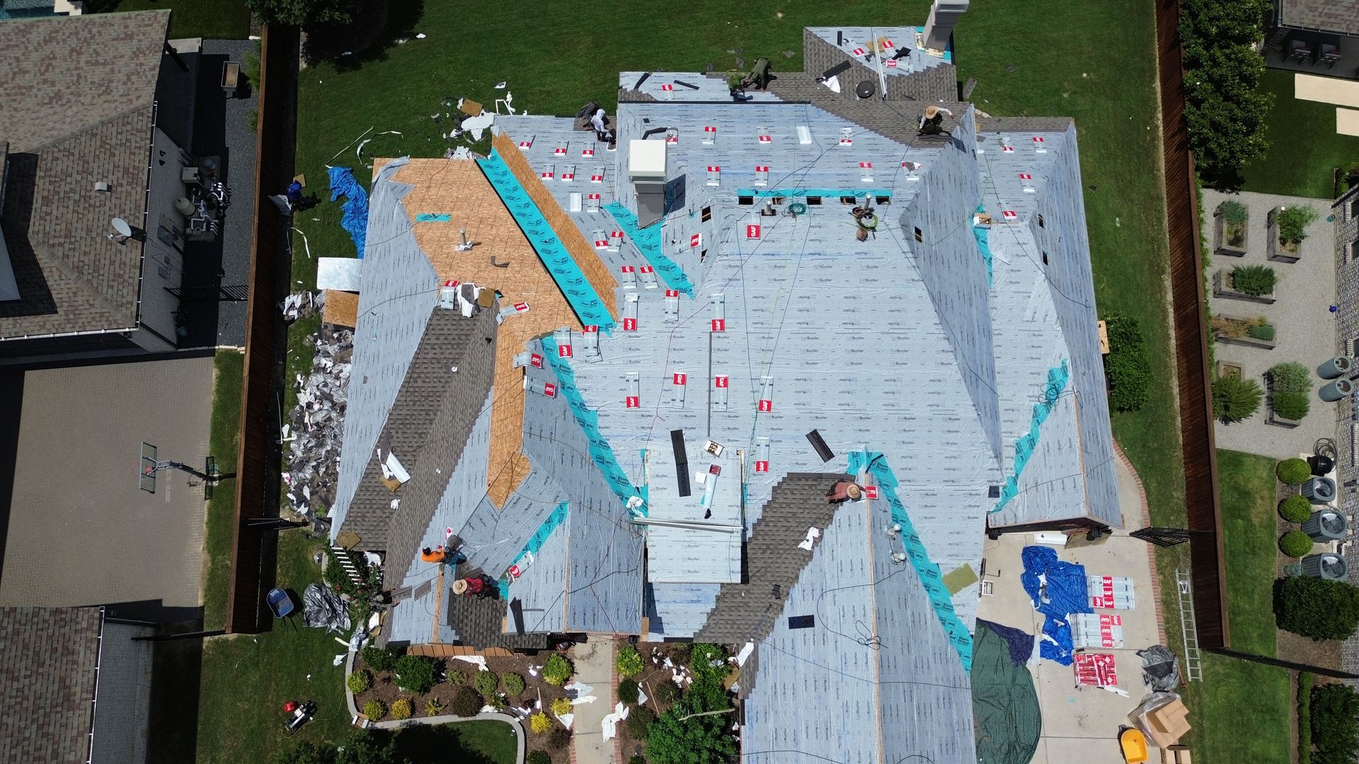 Aerial view of a house roof being repaired. Shingles partially removed. Green grass and surrounding houses visible.