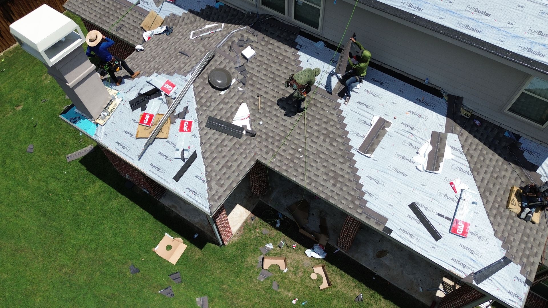 Roofers working on a house roof, with tools and materials visible. Green grass and blue sky background.