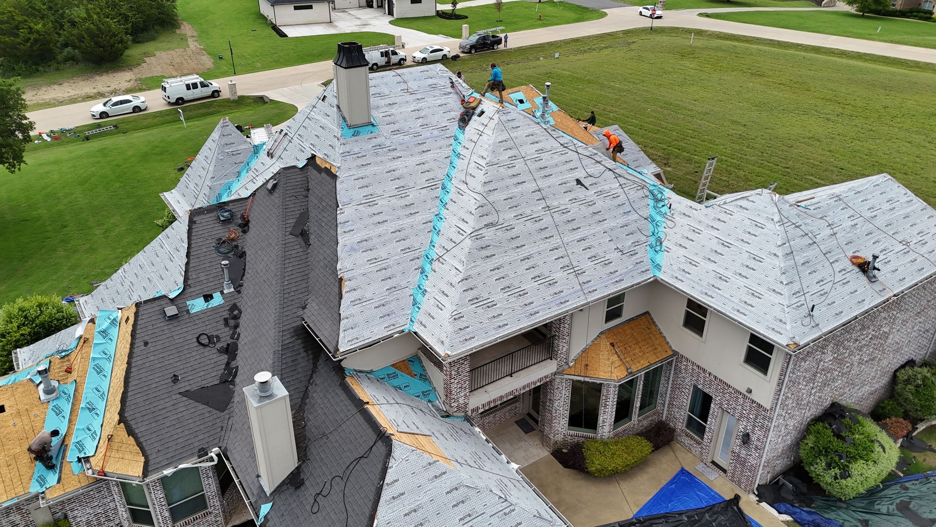 Roofing crew working on a large residential roof; various stages of completion, blue underlayment visible.