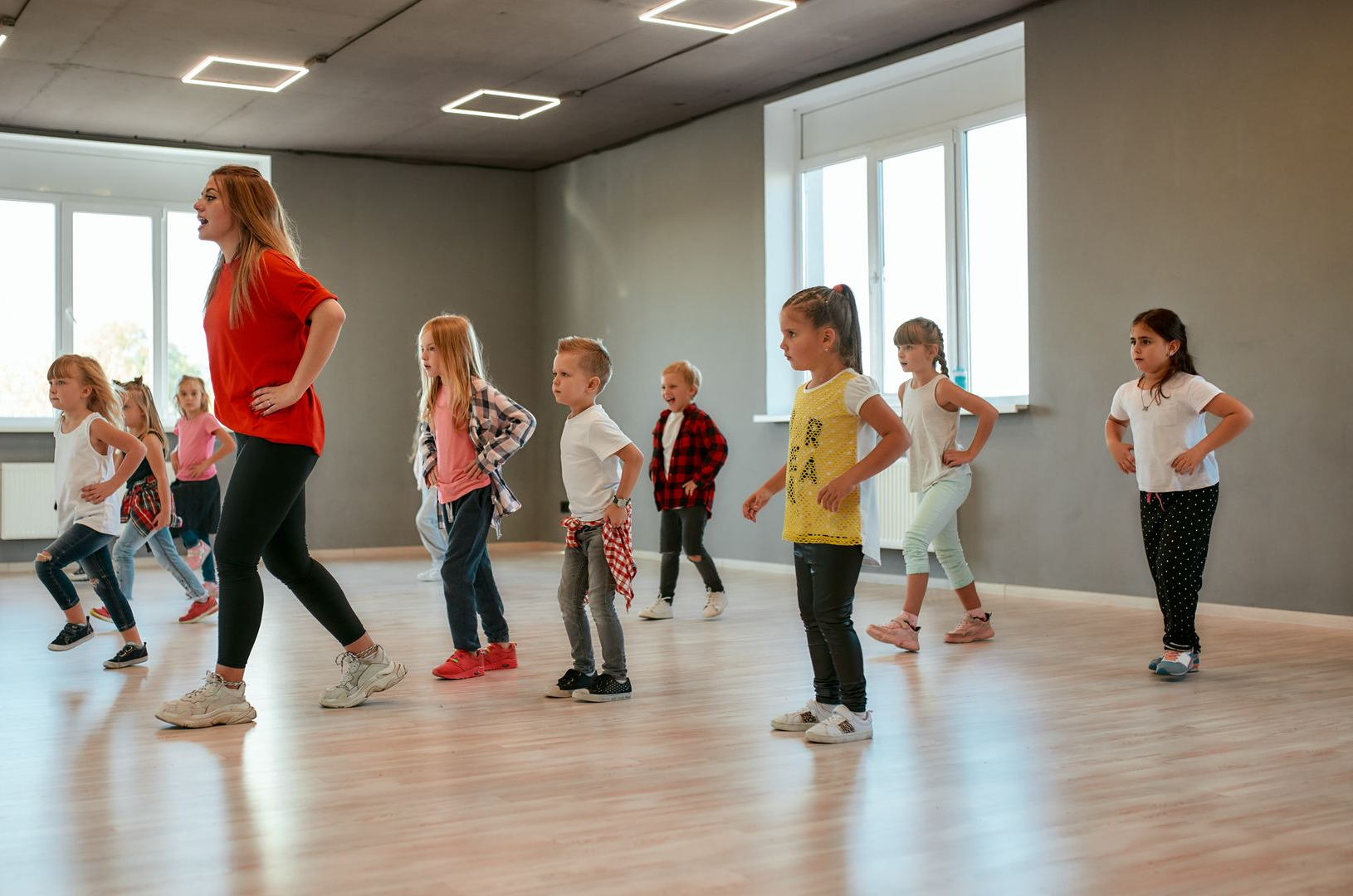 A teacher stands facing a group of students in a classroom with wooden desks, some of whom have their hands raised.